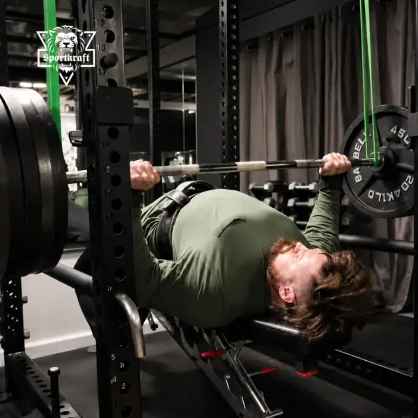 Urheilija tekee reverse band penkkipunnerrusta kuntosalilla power rack -telineessä vihreiden SportKraft Average vastuskumien varassa.