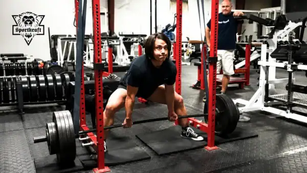 Voimanostaja tekee sumomaastavetoa kuntosalilla power rack -telineessä, jossa paksut mustat SportKraft Monster vastuskuminauhat on viritetty hangbar-kevennykseen.