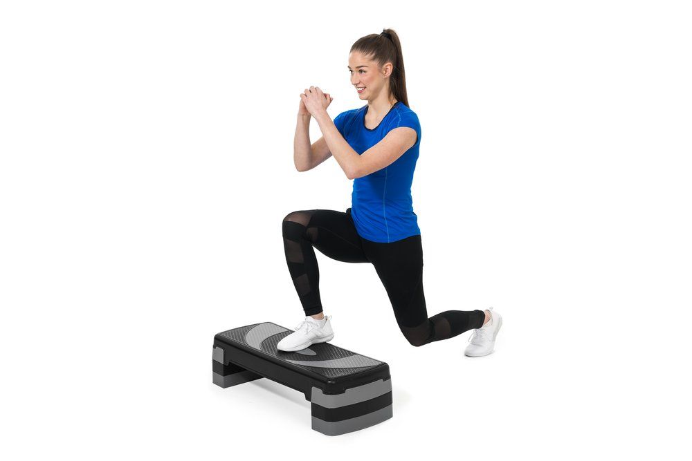 Nainen tekee askelkyykkyä steppilaudalla kotitreenissä, sininen paita ja mustat leggingsit.